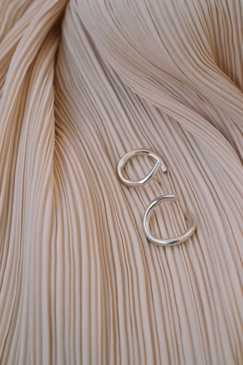 May include: Two silver-colored, open-ended hoop earrings. The earrings are set against a backdrop of pleated, beige fabric. The earrings have a simple, modern design, with a curved shape that wraps around the earlobe.
