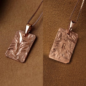 Puede incluir: Dos collares de oro con colgantes rectangulares. Un colgante tiene una superficie texturizada y el otro tiene la letra "R" grabada.