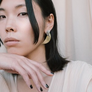 May include: A woman with dark hair wearing a light beige top and gold earrings. Her nails are painted black.