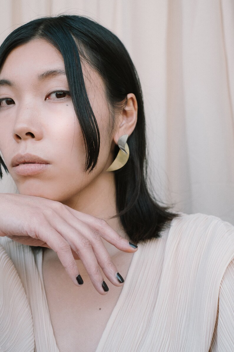 May include: A woman with dark hair wearing a light beige top and gold earrings. Her nails are painted black.