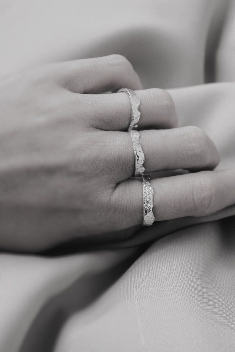 May include: Three silver rings with a textured, wavy design. The rings are stacked on a finger.