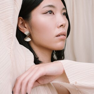 May include: A woman with dark hair wearing a light pink pleated blouse and silver earrings. She is looking off to the side with her hand resting on her shoulder.