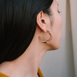 May include: A woman with long black hair wearing a gold hoop earring. The earring is textured and has a slightly curved shape.