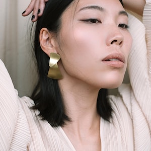 Puede incluir: Una mujer con cabello oscuro que lleva una blusa blanca plisada y pendientes geom&eacute;tricos dorados.
