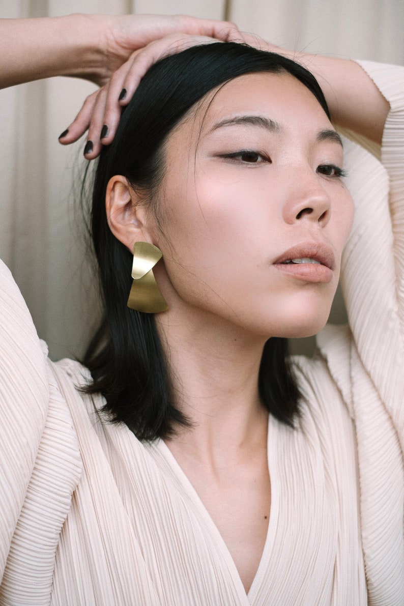 Puede incluir: Una mujer con cabello oscuro que lleva una blusa blanca plisada y pendientes geom&eacute;tricos dorados.