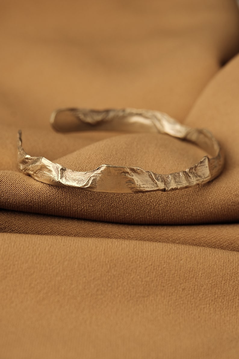 May include: A silver bracelet with a textured, mountain-like design. The bracelet is lying on a brown fabric surface.