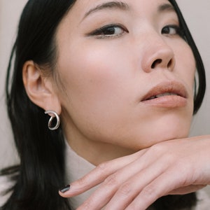 May include: A woman wearing a silver, open-hoop earring. The earring has a curved design. She is wearing a beige turtleneck. Her dark hair is styled straight. Her nails are painted dark. The image is a close-up.