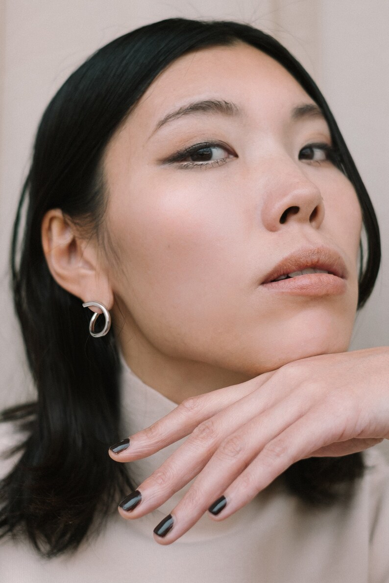 May include: A woman wearing a silver, open-hoop earring. The earring has a curved design. She is wearing a beige turtleneck. Her dark hair is styled straight. Her nails are painted dark. The image is a close-up.