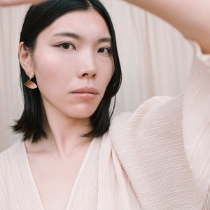 May include: A woman with short black hair wearing a cream-coloured, pleated blouse with a deep V-neck. She is wearing gold earrings and has her arm raised, partially obscuring her face.