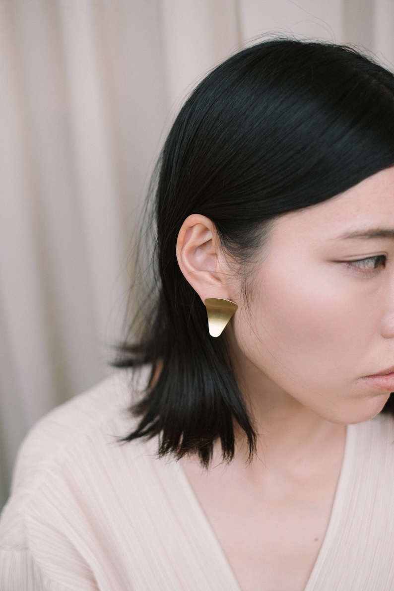 Puede incluir: Una mujer con el pelo negro corto lleva un par de pendientes triangulares de oro.