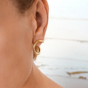 May include: Gold-colored spiral earrings. The earrings are a simple, minimalist design, with a smooth, polished finish. The earrings are shown on a person's ear, highlighting their shape and style.