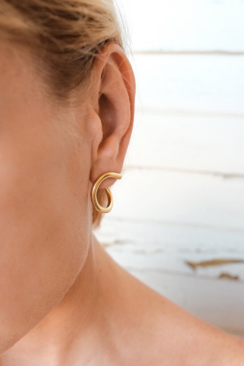 May include: Gold-colored spiral earrings. The earrings are a simple, minimalist design, with a smooth, polished finish. The earrings are shown on a person's ear, highlighting their shape and style.