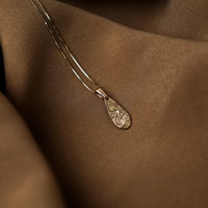 Peut inclure: Un collier d&eacute;licat en cha&icirc;ne dor&eacute;e avec un pendentif en forme de larme textur&eacute;. Le collier est pr&eacute;sent&eacute; sur un fond en tissu brun.