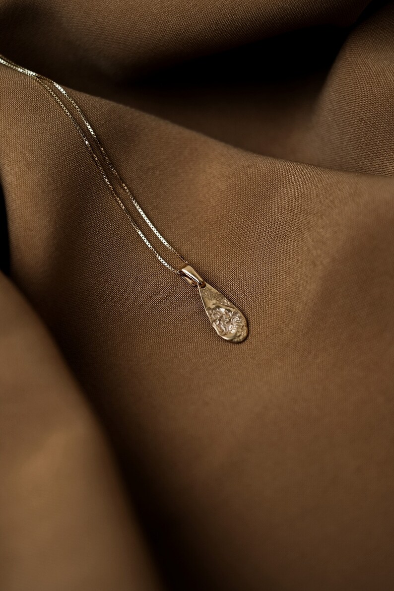 Peut inclure: Un collier d&eacute;licat en cha&icirc;ne dor&eacute;e avec un pendentif en forme de larme textur&eacute;. Le collier est pr&eacute;sent&eacute; sur un fond en tissu brun.