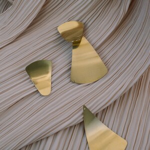 May include: Three gold-toned geometric earrings with a fan-shaped design. The earrings are on a light brown fabric background.