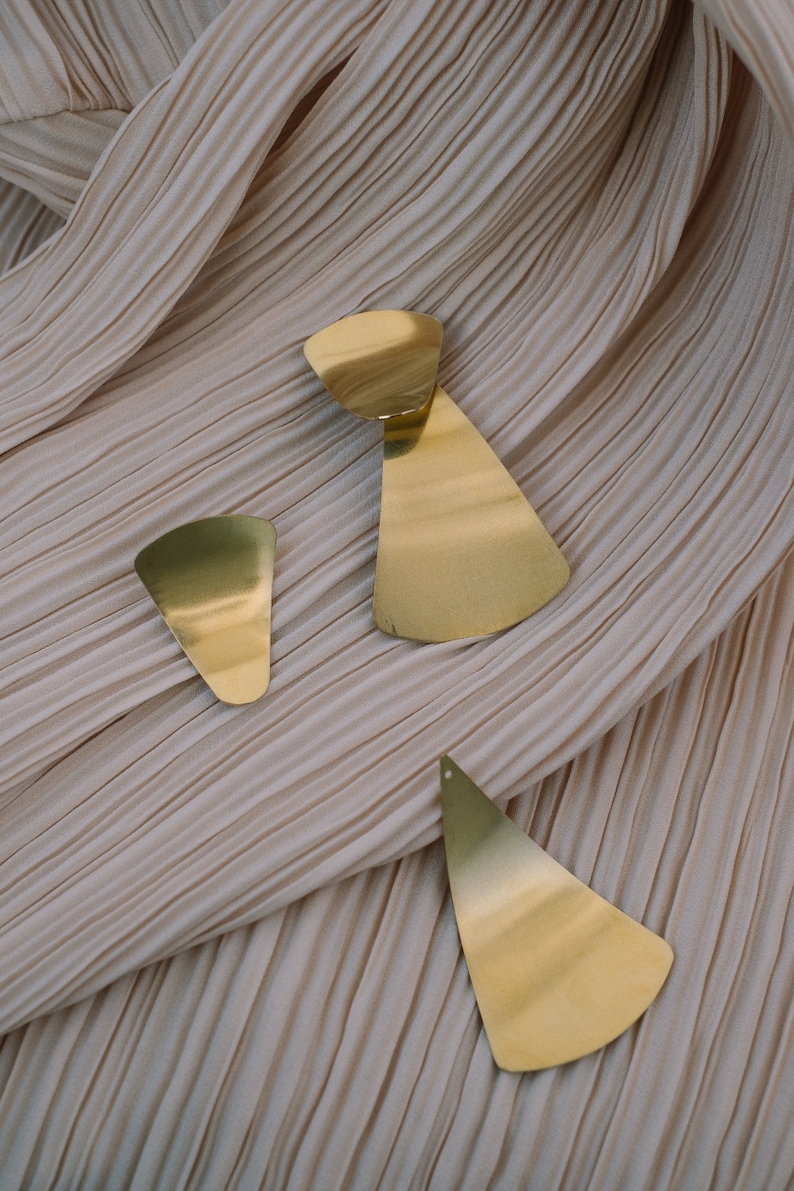 May include: Three gold-toned geometric earrings with a fan-shaped design. The earrings are on a light brown fabric background.