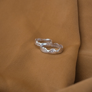May include: Two silver rings with a textured, mountain-like design. The rings are stacked on top of each other and are lying on a brown fabric surface.