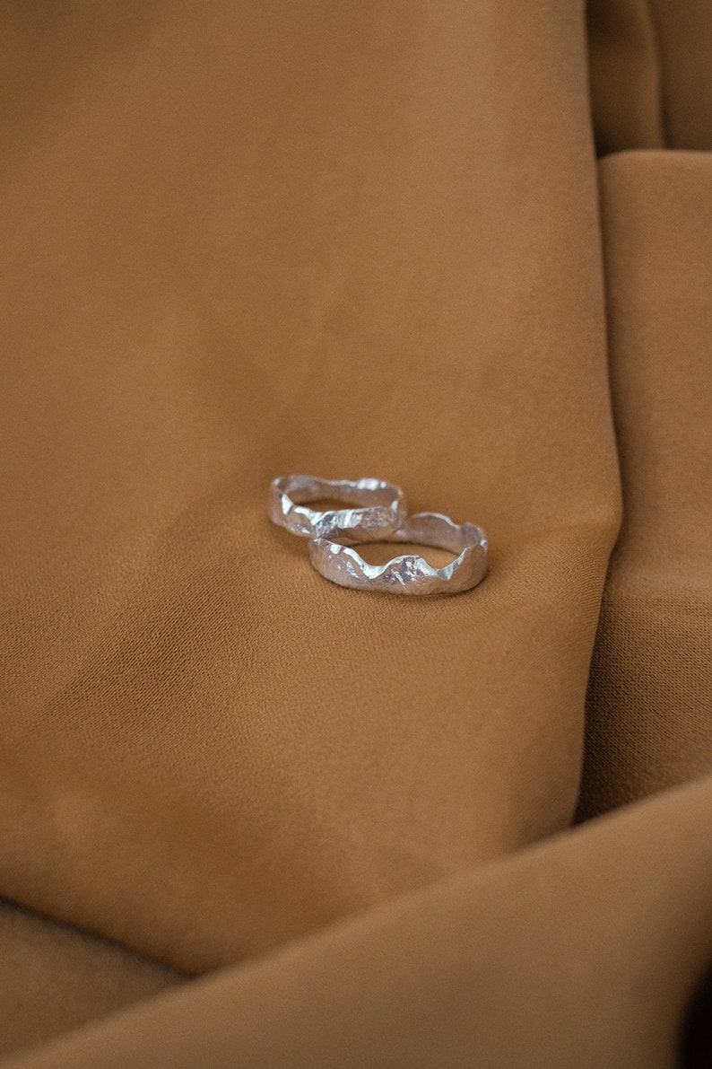 May include: Two silver rings with a textured, mountain-like design. The rings are stacked on top of each other and are lying on a brown fabric surface.