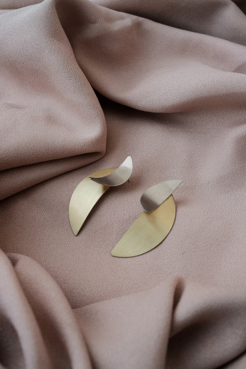 May include: A pair of gold and silver earrings with a geometric design. The earrings are on a light brown fabric background.