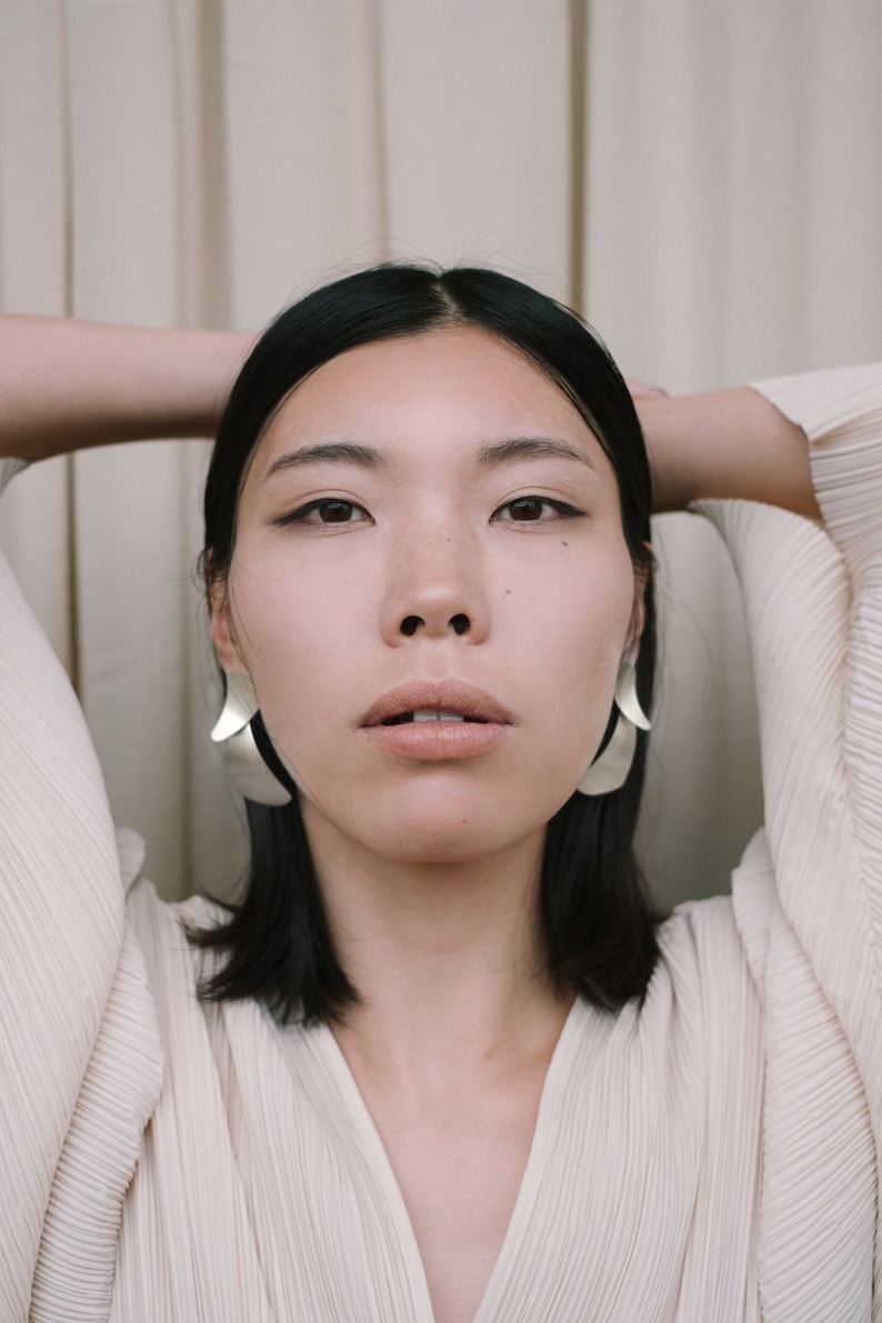 May include: A woman with dark hair wearing a white, pleated blouse and silver earrings. She is looking directly at the camera with her arms raised above her head.