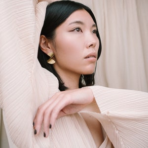 May include: A woman with dark hair wearing a light pink pleated blouse with long sleeves. She is wearing gold earrings with a geometric design.