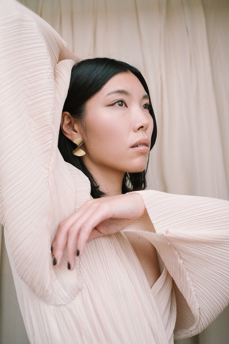 May include: A woman with dark hair wearing a light pink pleated blouse with long sleeves. She is wearing gold earrings with a geometric design.