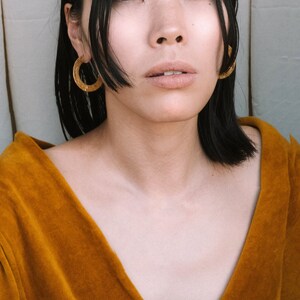 May include: A woman with black hair wearing a mustard yellow velvet top and gold hoop earrings.