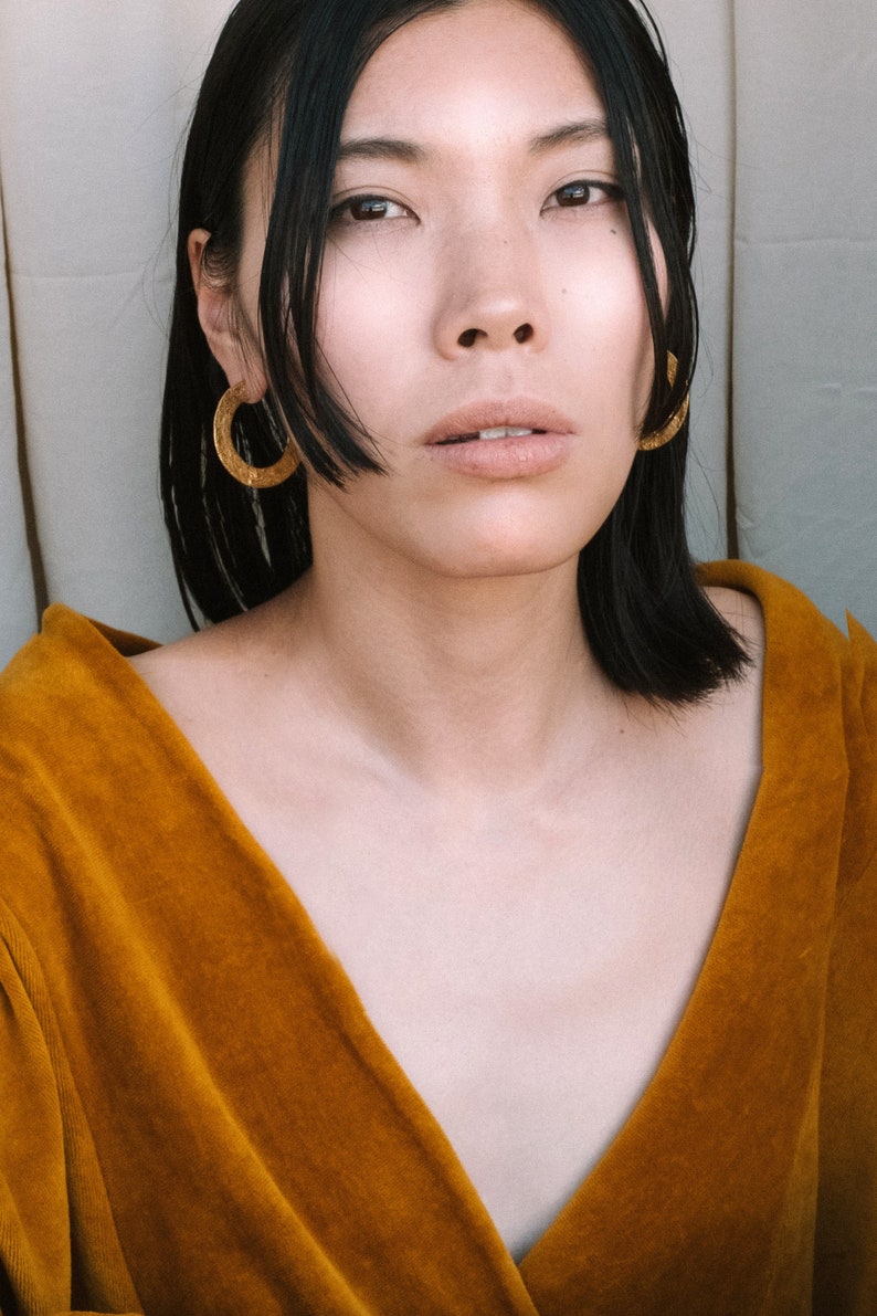 May include: A woman with black hair wearing a mustard yellow velvet top and gold hoop earrings.