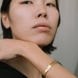 May include: A woman wearing a black shirt with a gold bracelet on her wrist. The bracelet has a textured design.