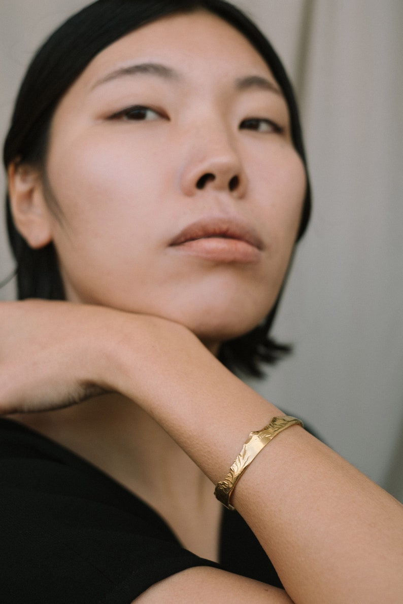 May include: A woman wearing a black shirt with a gold bracelet on her wrist. The bracelet has a textured design.