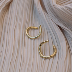 May include: Two gold-colored ear cuffs are displayed on a cream-colored, pleated fabric. The ear cuffs have a simple, curved design, with a small opening at the bottom. The fabric has a soft, flowing texture.