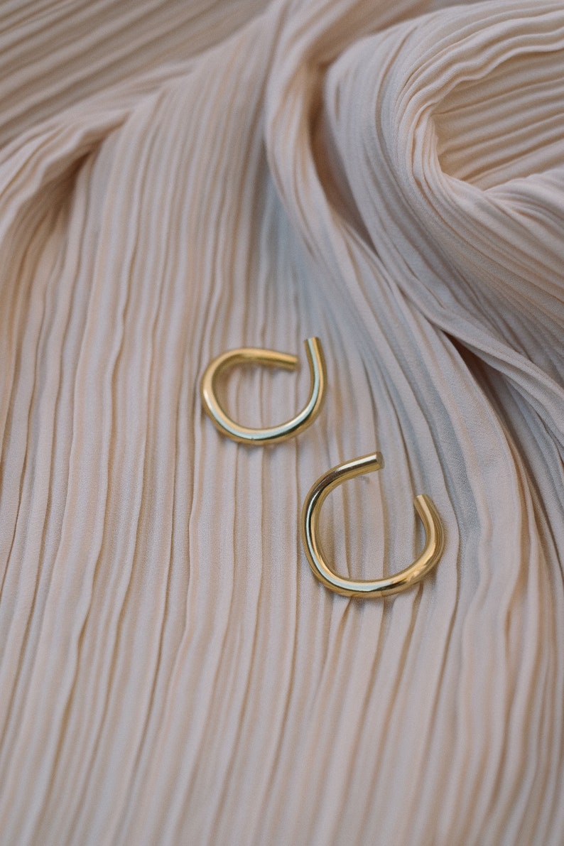 May include: Two gold-colored ear cuffs are displayed on a cream-colored, pleated fabric. The ear cuffs have a simple, curved design, with a small opening at the bottom. The fabric has a soft, flowing texture.