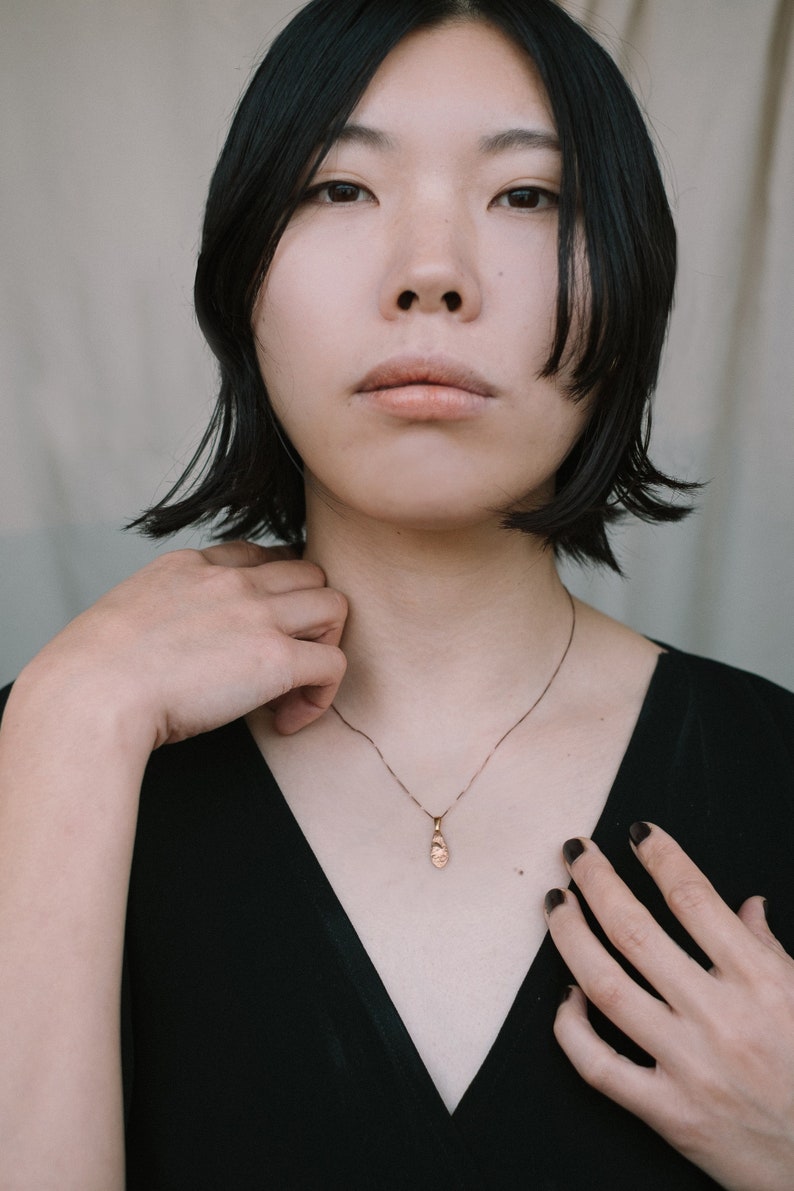 Peut inclure: Une femme aux cheveux noirs courts portant un haut noir &agrave; col en V et un collier en cha&icirc;ne dor&eacute;e avec un petit pendentif.