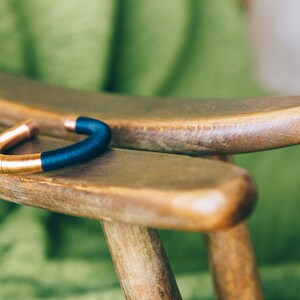 Pu&ograve; includere: Un braccialetto in rame e blu con un design semplice. Il braccialetto &egrave; appoggiato su una sedia di legno.