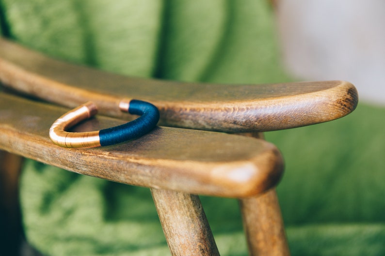 Pu&ograve; includere: Un braccialetto in rame e blu con un design semplice. Il braccialetto &egrave; appoggiato su una sedia di legno.