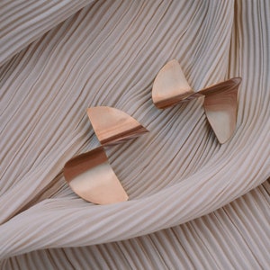May include: Two pairs of gold-toned geometric earrings with a twisted design. The earrings are on a beige fabric background.
