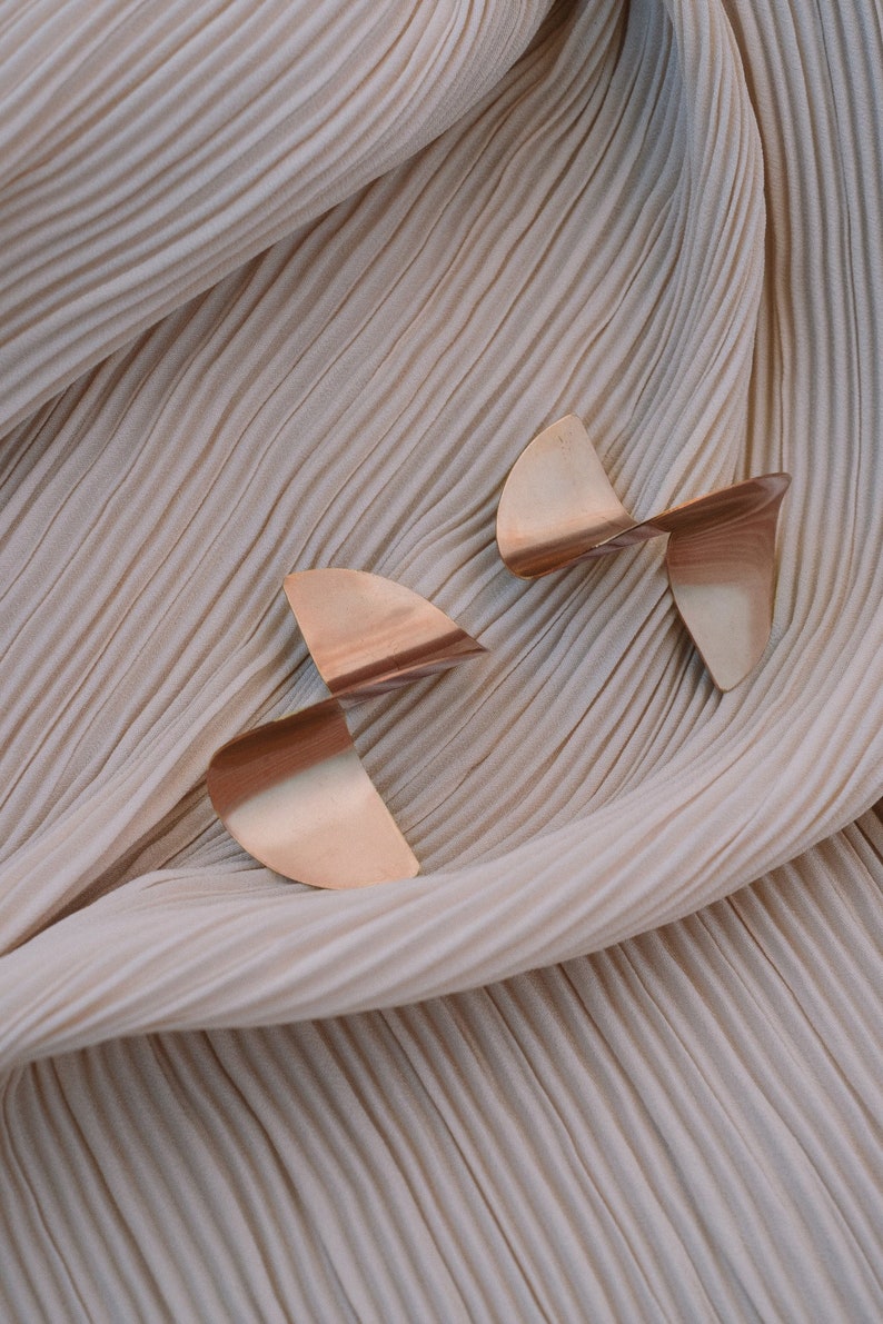 May include: Two pairs of gold-toned geometric earrings with a twisted design. The earrings are on a beige fabric background.