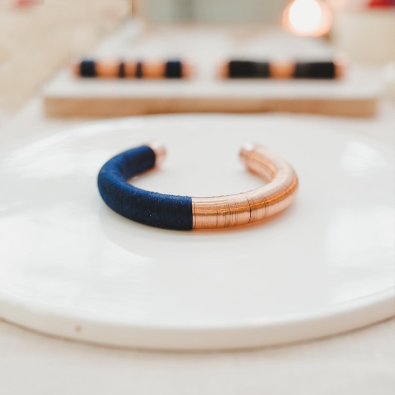 Pu&ograve; includere: Un braccialetto rotondo in rame e blu navy. Il braccialetto &egrave; realizzato con filo di rame avvolto attorno a una fascia di tessuto blu.