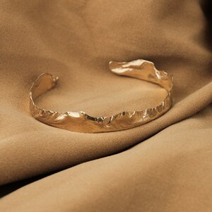 May include: A gold-toned, textured metal bracelet with a unique, organic shape. The bracelet is lying on a brown fabric surface.