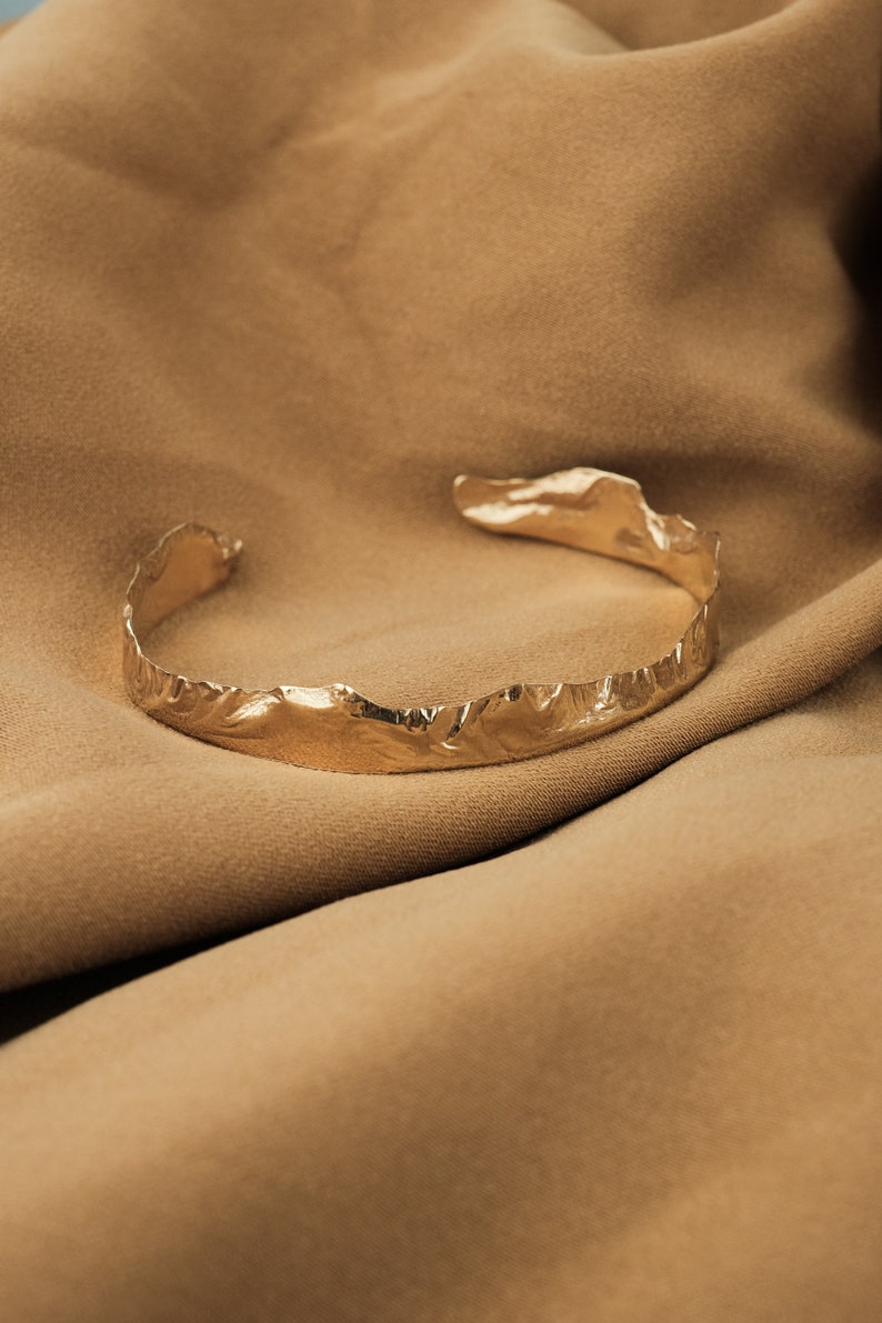 May include: A gold-toned, textured metal bracelet with a unique, organic shape. The bracelet is lying on a brown fabric surface.