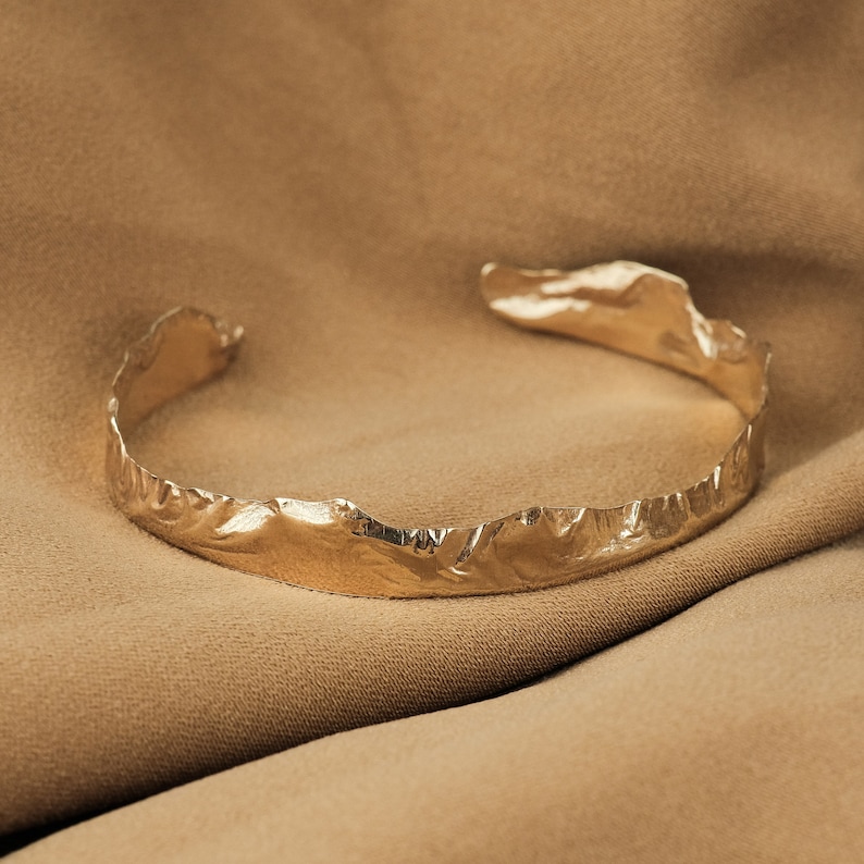 May include: A gold-toned metal bracelet with a textured, organic design. The bracelet is on a brown fabric background.