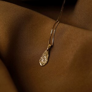 Peut inclure: Un collier en or avec un pendentif en forme de larme. Le pendentif a une surface textur&eacute;e et est suspendu sur un fond en tissu brun.