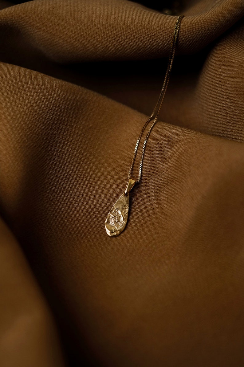 Peut inclure: Un collier en or avec un pendentif en forme de larme. Le pendentif a une surface textur&eacute;e et est suspendu sur un fond en tissu brun.