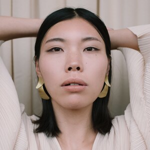 May include: A woman with dark hair wearing a white, pleated blouse and gold earrings. She is looking directly at the camera.