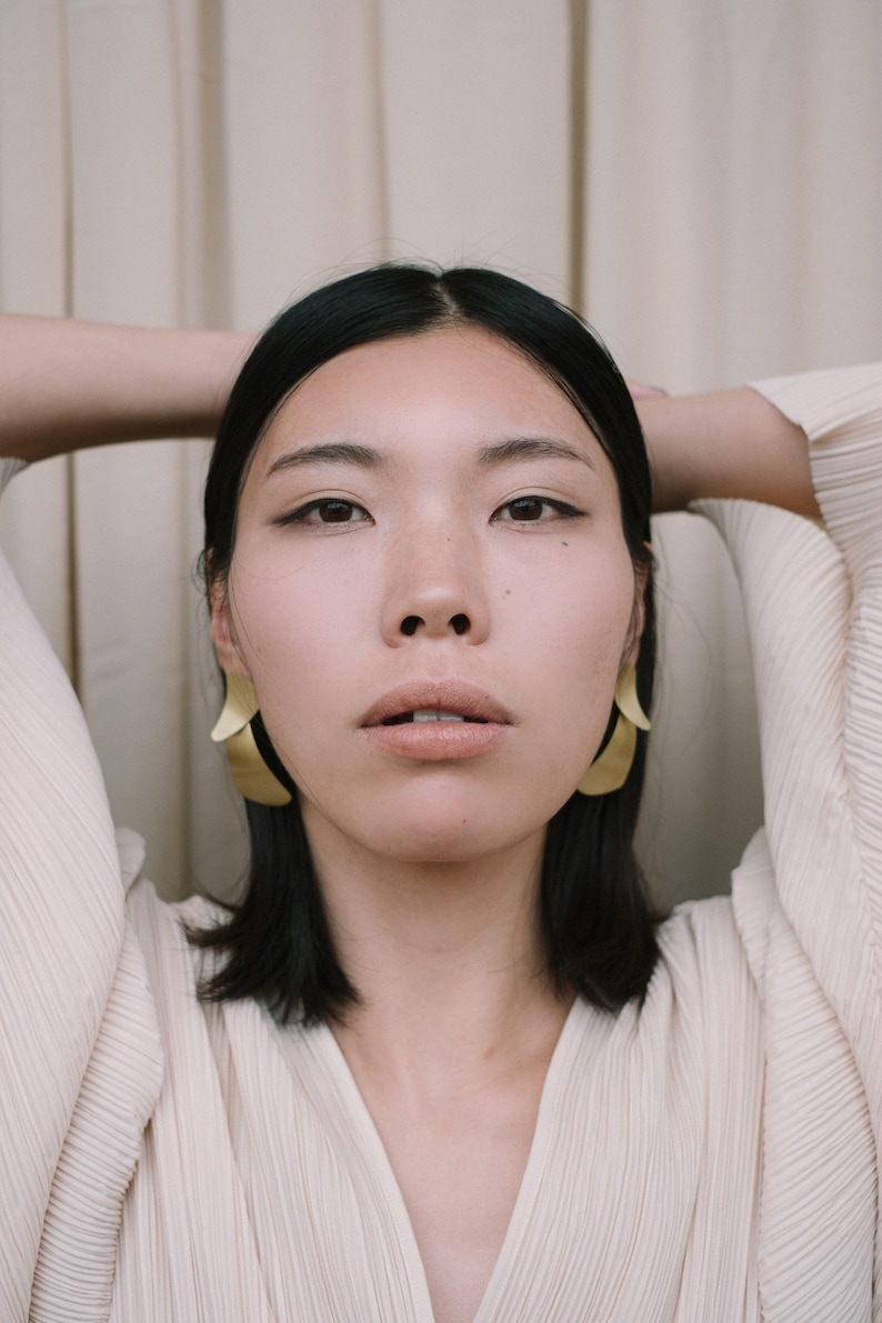 May include: A woman with dark hair wearing a white, pleated blouse and gold earrings. She is looking directly at the camera.