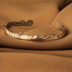 Könnte beinhalten: Ein goldfarbenes Metallarmband mit einem strukturierten, organischen Design. Das Armband ist offen und hat eine raue, unebene Oberfläche.