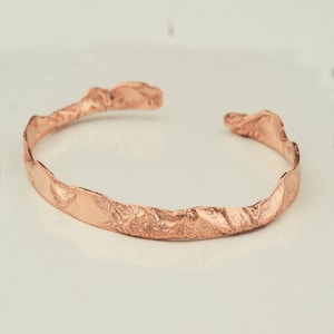 Puede incluir: Un brazalete de oro rosa con un diseño texturizado y orgánico. El brazalete está hecho de metal y tiene un acabado liso y pulido.