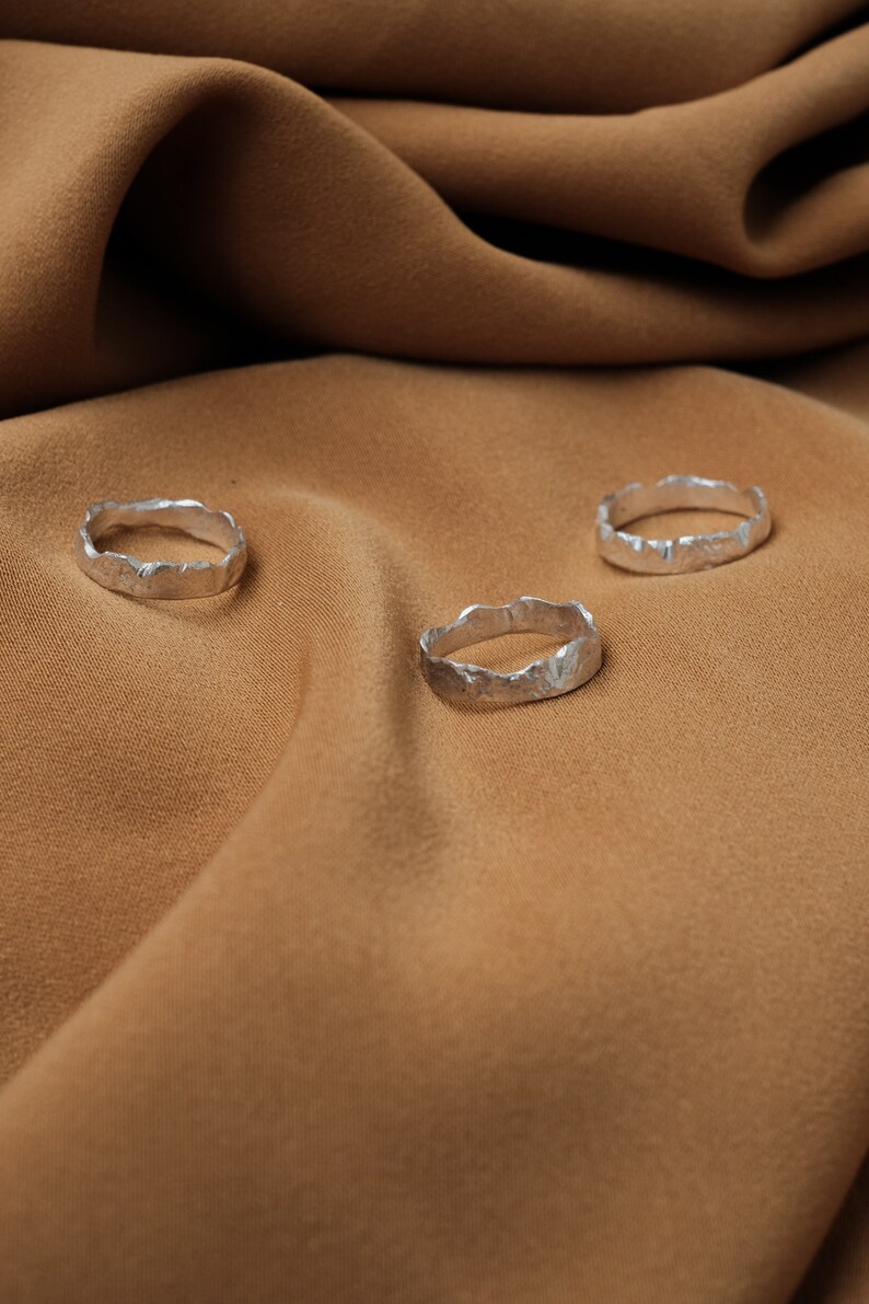 May include: Three silver rings with a textured, organic design, arranged on a brown fabric background.