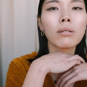 May include: A portrait featuring a person wearing gold hoop earrings. The person is wearing a mustard-colored velvet top. The person's hands are gently clasped near the face. The background is a neutral color.