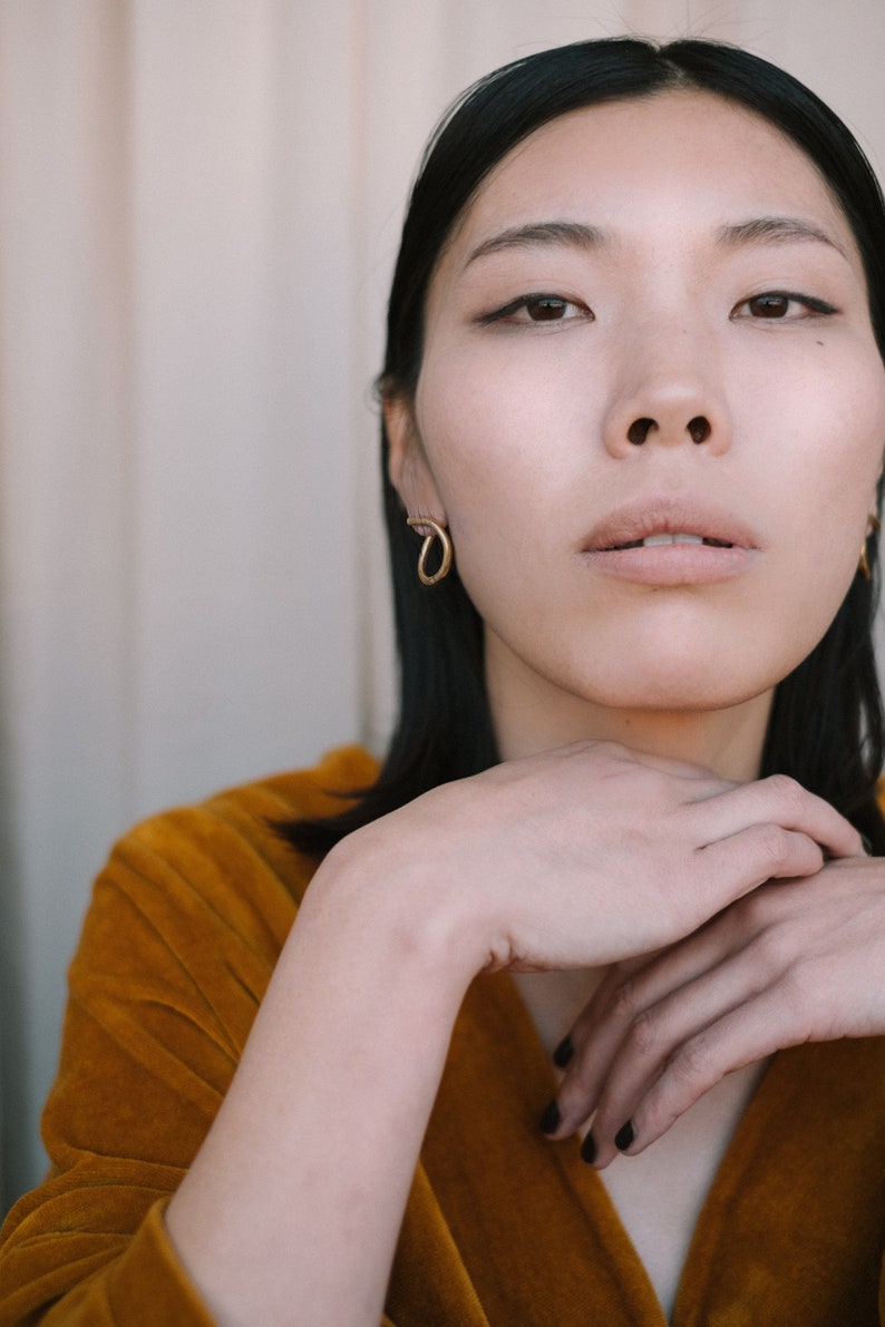 May include: A portrait featuring a person wearing gold hoop earrings. The person is wearing a mustard-colored velvet top. The person's hands are gently clasped near the face. The background is a neutral color.